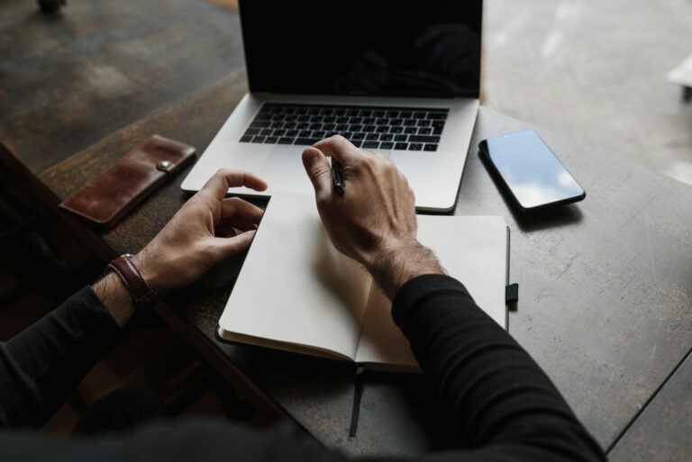 Overhead view of hands writing in a notebook beside a laptop and smartphone on a wooden desk, considering digital vs paper journaling